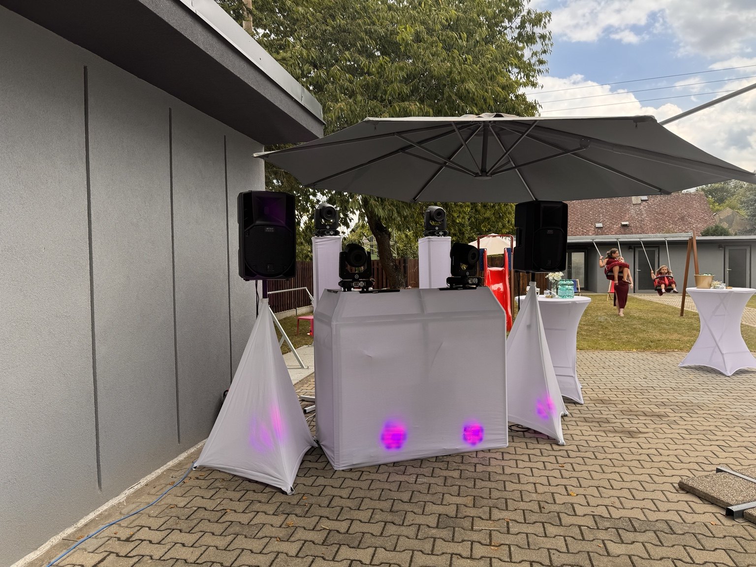Sestava DJ Wendys na venkovní akci: dvě reprosoustavy RCF na stojanech, čtyři pohyblivé světelné hlavy a DJ pult s bílým čelem pod slunečníkem